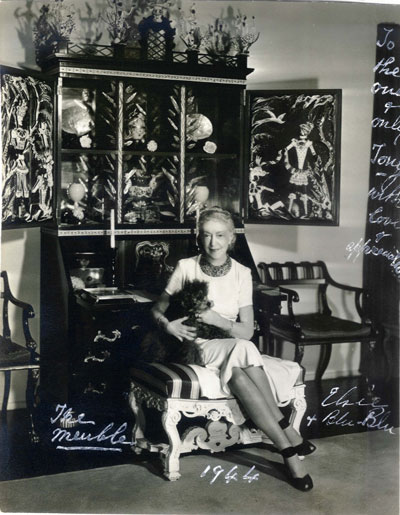 Elsie de Wolfe with her blue-dyed hair and blue-dyed poodle (named Blu-Blu, of course) dressed by Mainbocher, wearing her sapphire and diamond bib necklace by Madame Belperon, seated in front of the secretary desk that she commissioned from Tony Duquette, c. 1944.